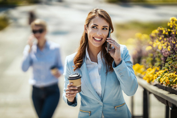Νεαρή χαμογελαστή επιχειρηματίας περπατά μπροστά από το γραφείο και μιλάει στο smartphone κατά τη διάρκεια του διαλείμματος καφέ. - Φωτογραφία, εικόνα