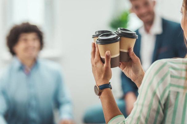 Молодая женщина несет одноразовые кофейные чашки своим улыбающимся коллегам во время перерыва на кофе в офисе. - Фото, изображение