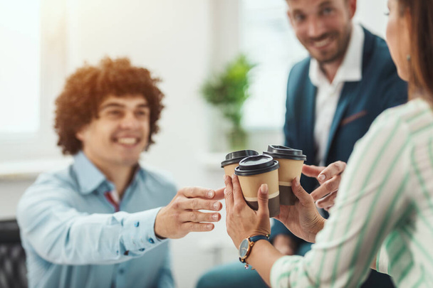 Молодая женщина несет одноразовые кофейные чашки своим улыбающимся коллегам во время перерыва на кофе в офисе. - Фото, изображение