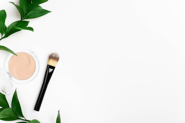 Face powder in round case and make up brush top view with green leaf background. Fashion cosmetic, natural makeup product composition. Open compact foundation cream. Organic facial concealer powder. - Photo, Image