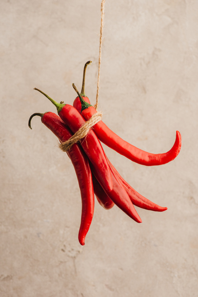 ένα μάτσο κόκκινες πιπεριές τσίλι δεμένες με σχοινί κρεμασμένες σε μπεζ φόντο από τσιμέντο - Φωτογραφία, εικόνα