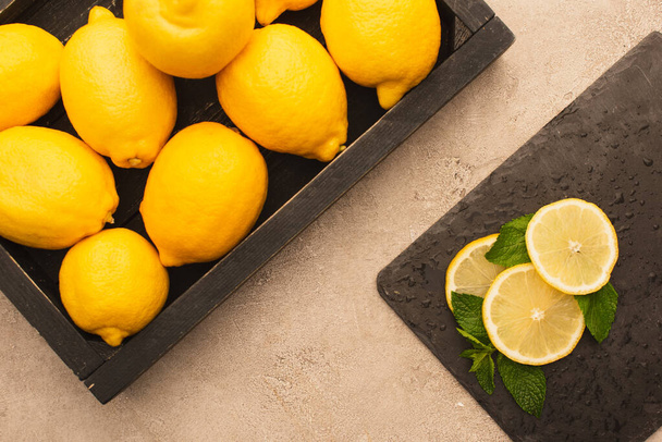 top view of whole yellow lemons in box and sliced on board with mint green leaves on beige concrete surface - Foto, imagen