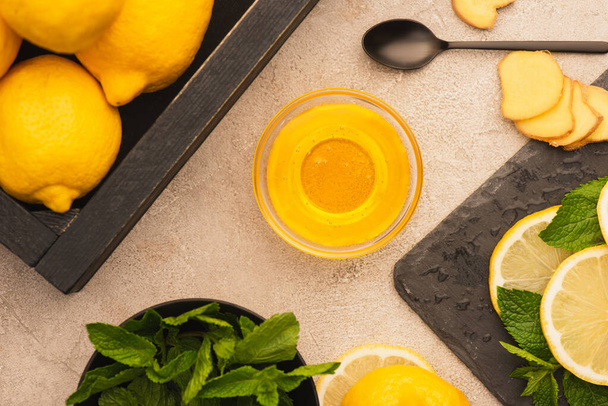top view of yellow lemons, mint green leaves, ginger root slices and honey on beige concrete surface - Fotografie, Obrázek