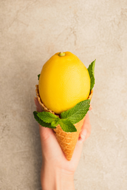 cropped view of woman holding waffle cone with yellow lemon with mint green leaves on beige concrete surface - Photo, Image