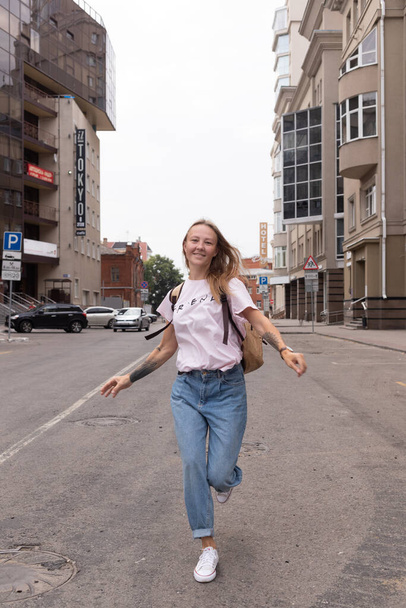 a beautiful smiling girl in a white t-shirt and jeans poses on the streets of the city. she has long hair and no makeup. the architecture of the old city is a backdrop around it - Foto, Bild