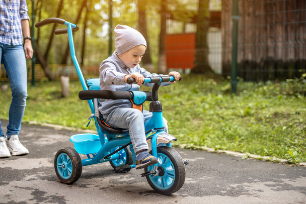 Little boy is learning to ride a children's tricycle while walking with his mother. Concept of learning to ride a bike and spend active time with your family. - 写真・画像