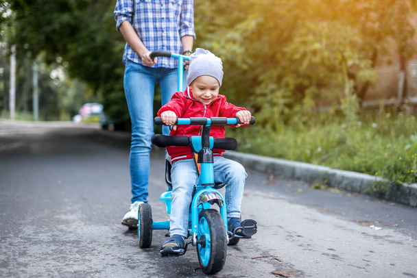 Young mother is pushing a child's tricycle with a toddler boy on a walk. Concept of learning to ride a bike and having fun with your family. - Фото, зображення