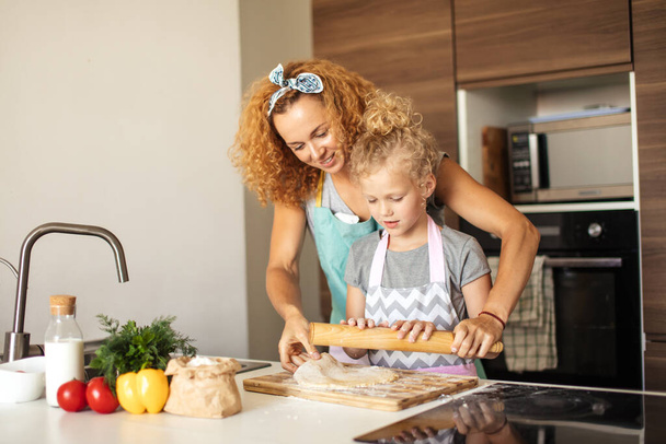 Beautiful young mom and her cute little daughter rolling out dough at home. - 写真・画像