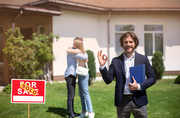 Smiling realtor showing okay gesture and millennial couple embracing near their new property, copy space - Photo, Image