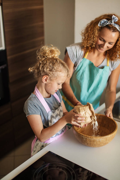 Little girl and her mother sprinkling flour into a bowl and smiling while baking - Foto, Imagem