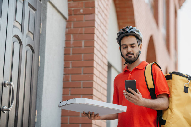 Fast delivery of pizza by courier in city. Serious man in helmet and bag, holds cardboard box with food and watches at smartphone - Фото, изображение