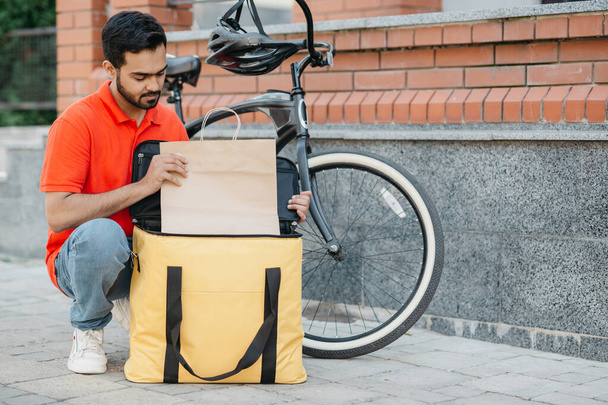 Courier and package for the client. Busy guy courier in uniform with bicycle puts goods in bag - Φωτογραφία, εικόνα