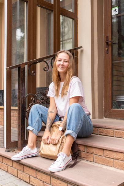 a beautiful smiling girl in a white t-shirt and jeans poses on the streets of the city. she has long hair and no makeup. the architecture of the old city is a backdrop around it - Valokuva, kuva