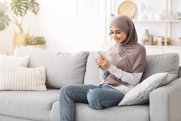 Moderna ragazza musulmana che utilizza smartphone a casa mentre si rilassa sul divano - Foto, immagini