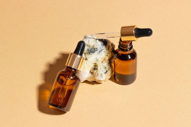 Natural cosmetics in glass bottles with a dropper stand next to a stone on a beige background with bright sunlight. The concept of natural cosmetics, natural essential oil - Fotó, kép