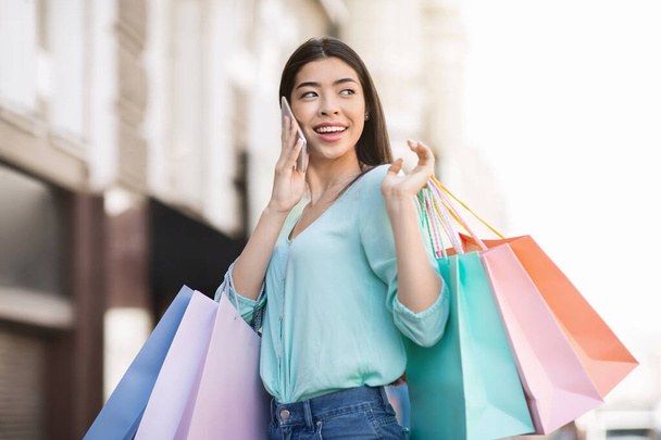 Feliz chica asiática caminando con muchas bolsas de compras y hablando en el teléfono celular - Foto, imagen