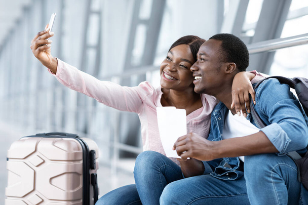 Путевые воспоминания. Веселая черная пара делает селфи на смартфоне в аэропорту - Фото, изображение