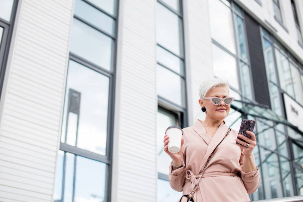 businesswoman using mobile phone and holding coffee cup against urban background - Foto, Imagem