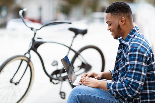 czarny człowiek za pomocą laptopa, siedzi w mieście z rower - Zdjęcie, obraz