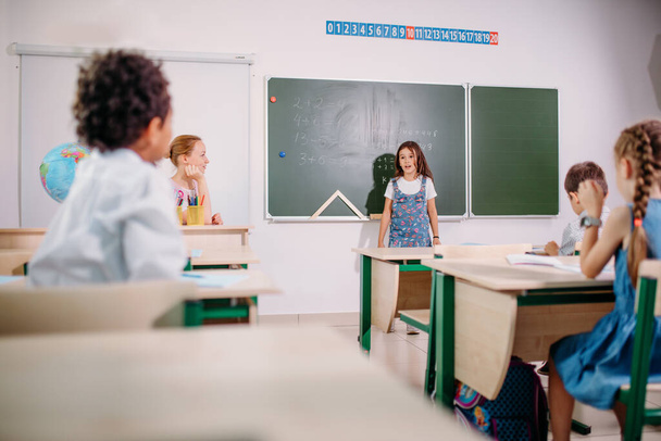 Schoolchild with teacher in classroom. Girl near chalkboard show her homework - 写真・画像