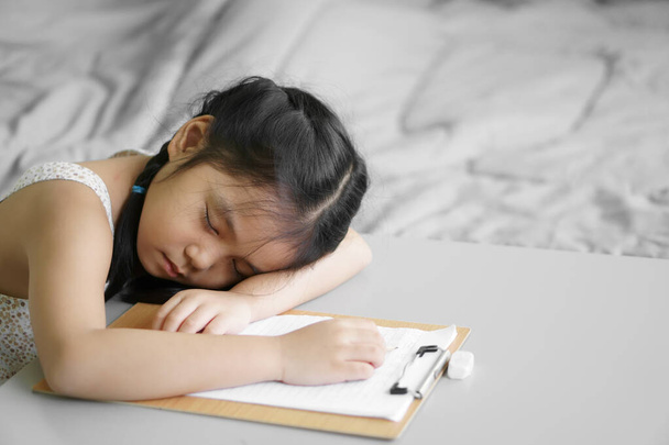азиатский ребенок студент или ребенок девочка пишет или рисовать и делать домашнюю работу или учиться из дома и обучение писать на бумаге, но засыпать или спать отдых на столе, оставаться в безопасности и учеба в школе онлайн в спальне - Фото, изображение