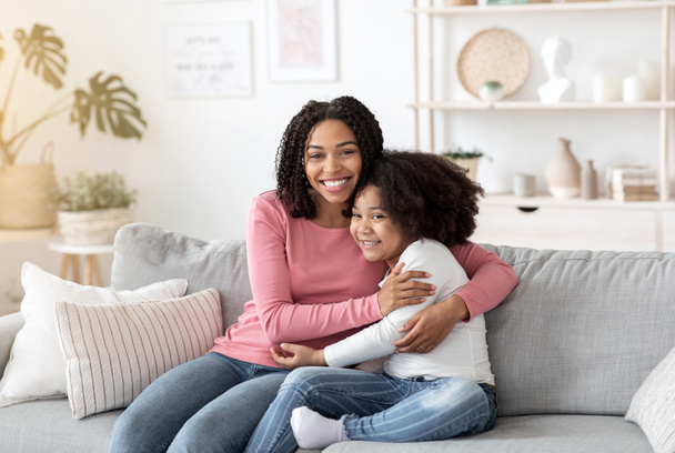 Happy Black Mother Embracing Her Cute Little Daughter On Couch At Home - Foto, Imagen