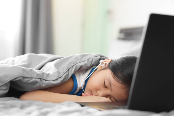 Aasian lapsi opiskelija tai lapsi tyttö käyttää kuuloke tai kuulokkeet opiskella verkossa tai työskennellä kotoa tietokoneen kannettavan tai pelata kannettavaa tietokonetta ja nukahtaa sängylle peruskoulun oppiminen ja pysyä turvassa - Valokuva, kuva