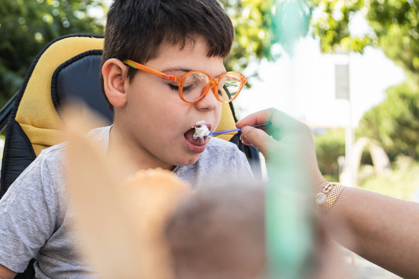 postižené dítě na invalidním vozíku jí zmrzlinu lžící. Ženská ruka mu podává jídlo. nosí oranžové brýle a šedé tričko. vnější - Fotografie, Obrázek
