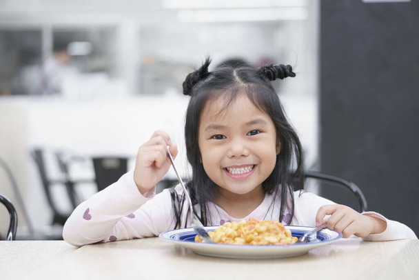 Niño asiático lindo o niña disfrutar de comer macarrones de pollo para la comida italiana y apetitoso en la mesa con cuchara de espera y sonriente dientes blancos a feliz para el desayuno o el almuerzo en el restaurante o patio de comidas - Foto, Imagen
