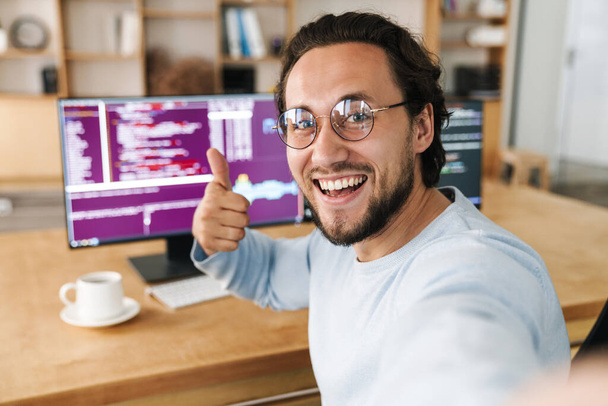 Image of unshaven excited programmer man showing thumb up while taking selfie photo in office - Фото, изображение