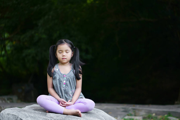 Ázsiai gyermek aranyos vagy gyerek lány szoros szem éberség meditáló lélegzet és ülni kő vagy szikla a béke és pihenés vagy jóga a természet zöld kert és fa a templom parkban, vagy meditálni templom szabadban - Fotó, kép