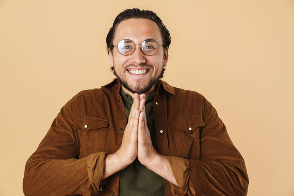 Image of handsome happy man in eyeglasses smiling and holding palms together isolated over beige background - Foto, Bild