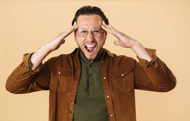 Beeld van jonge ongelukkige man met hoofdpijn in bril schreeuwen op camera geïsoleerd over beige achtergrond - Foto, afbeelding