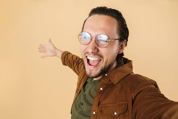 Image of young delighted man winking and laughing while taking selfie photo isolated over beige background - Foto, afbeelding