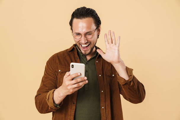 Image of excited bristle man in eyeglasses waving hand and using cellphone isolated over beige background - Фото, зображення