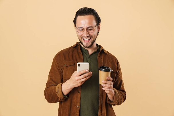 Image of cheerful bristle man drinking coffee takeaway and using cellphone isolated over beige background - 写真・画像