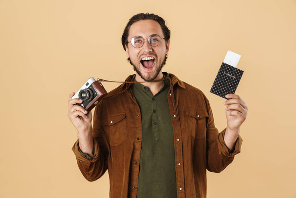 Image of delighted handsome man in eyeglasses holding retro camera and passport isolated over beige background - Zdjęcie, obraz