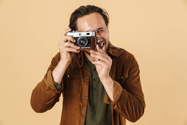 Image of laughing caucasian man taking photo on retro camera isolated over beige background - 写真・画像