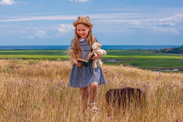 Child girl in a straw hat and dress staying near vintage suitcase and reading a book. Cute kid with soft dog toy looking at notebook in hands nature lanscape background. Adventure concept retro style. - Foto, immagini