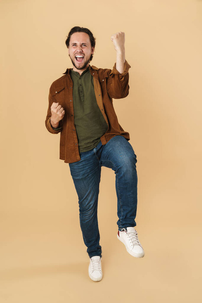 Image of excited man screaming and making winner gesture on camera isolated over beige background - Fotó, kép