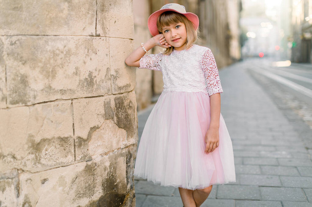 Cute little girl in fashionable pink hat and dress posing to camera outdoors in the city street, standing near old stone wall of ancient building in European city - Photo, image