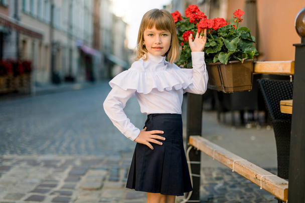 Closeup of little pretty blond Caucasian girl touching red flower pots decorations on summer day in old ancient European city - Zdjęcie, obraz