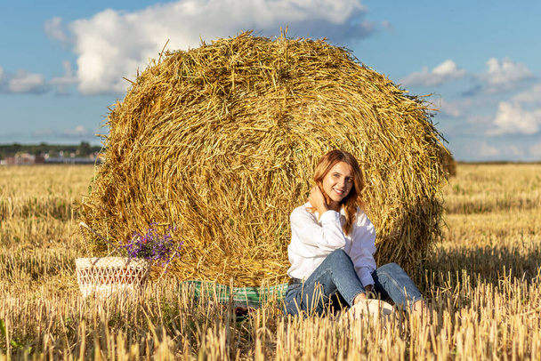 Beautiful young woman in a white shirt and jeans resting in a field near a hay bale. - Photo, Image