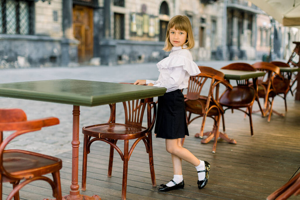 Pretty little girl in business elegant clothes, standing at the table of a street cafe with ancient city buildings on the blurred background - 写真・画像