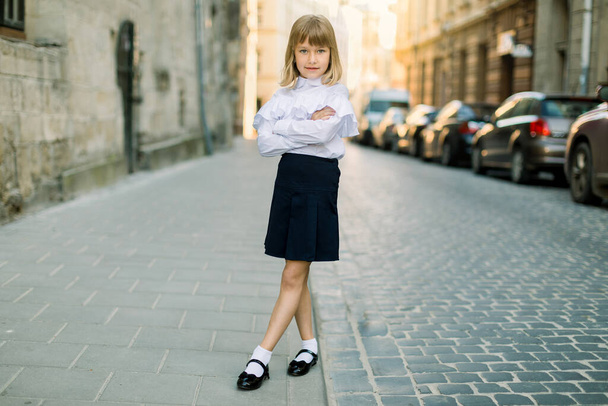 Full length portrait of beautiful little blond girl outdoors at old city street in Lviv, Ukraine. Fashion strret portrait of pretty girl in the city - Фото, изображение