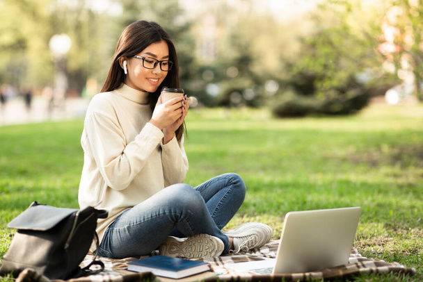 Девушка пьет кофе, пользуется ноутбуком, сидит в парке - Фото, изображение