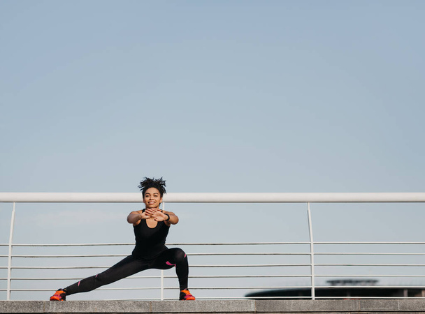 Frauen bewegen sich für eine gute Gesundheit. Glückliche junge Afrikanerin in Sportbekleidung mit Fitness-Tracker bei Beinübungen - Foto, Bild