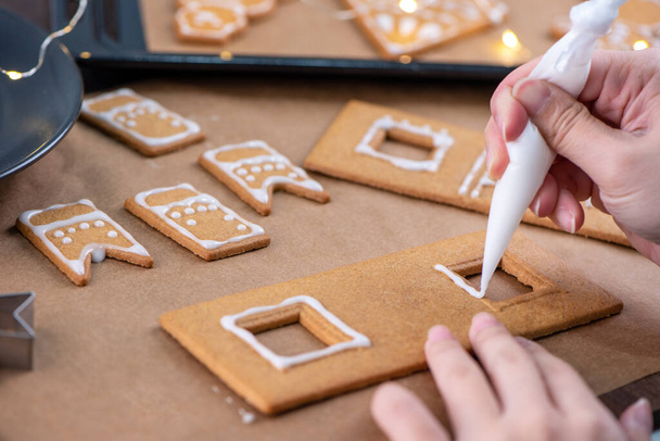 Young woman is decorating Christmas Gingerbread House cookies biscuit at home with frosting topping in icing bag, close up, lifestyle. - Photo, Image