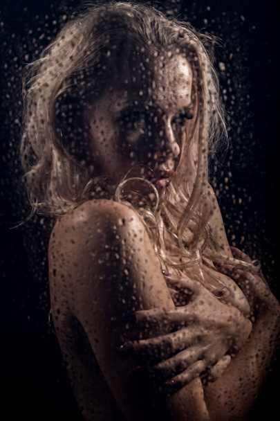 beautyful blonde woman behind wet glass on a dark background, woman takes a shower - Photo, Image