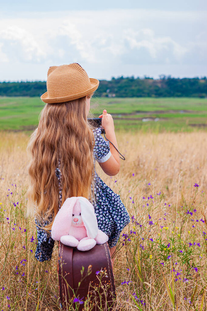 Dziewczynka z długimi włosami w słomkowym kapeluszu i sukience siedzi na zabytkowej walizce i robi zdjęcie. Słodki dzieciak z miękką zabawką patrząc na tle lanscape natury. Pojęcie podróży przygodowej w stylu retro - Zdjęcie, obraz